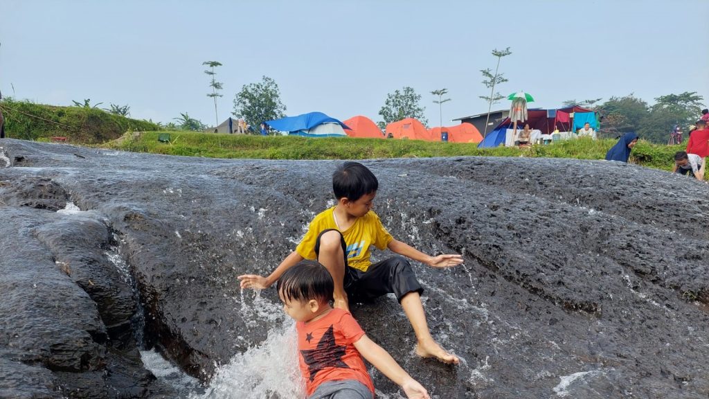 Area perkemahan Sisi Cai Camping Ground Sentul di pinggir sungai dengan tenda dan pepohonan hijau.